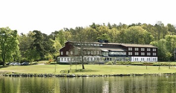A large building with multiple windows and a covered porch, situated next to a body of water and surrounded by trees.