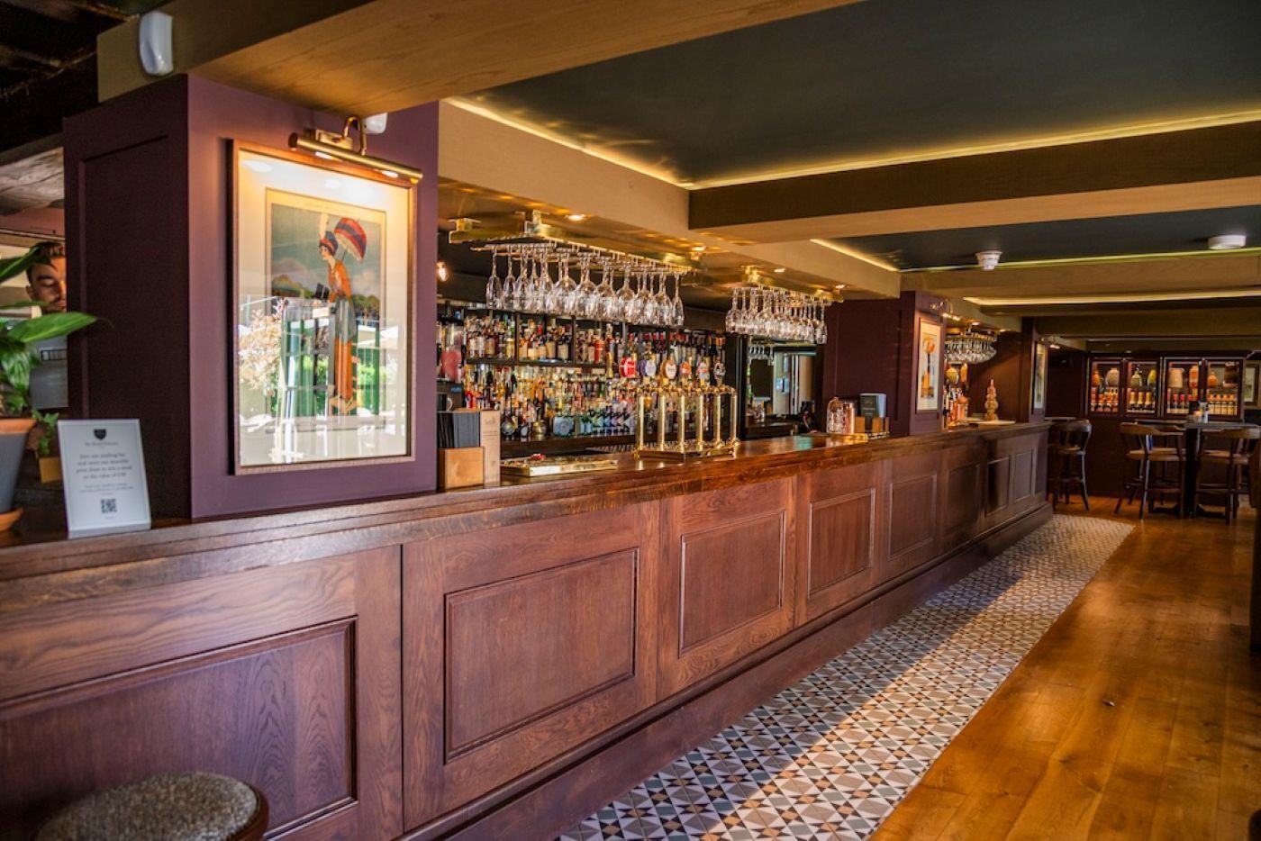 A bar area with a wooden counter, shelves of bottles, and hanging glasses.