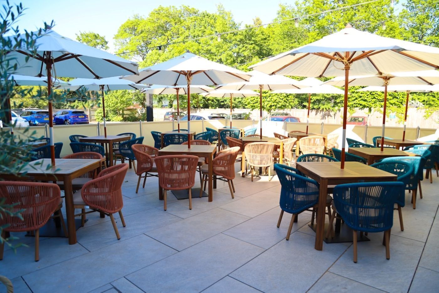 An outdoor patio with tables and chairs, umbrellas, and a parking area in the background.
