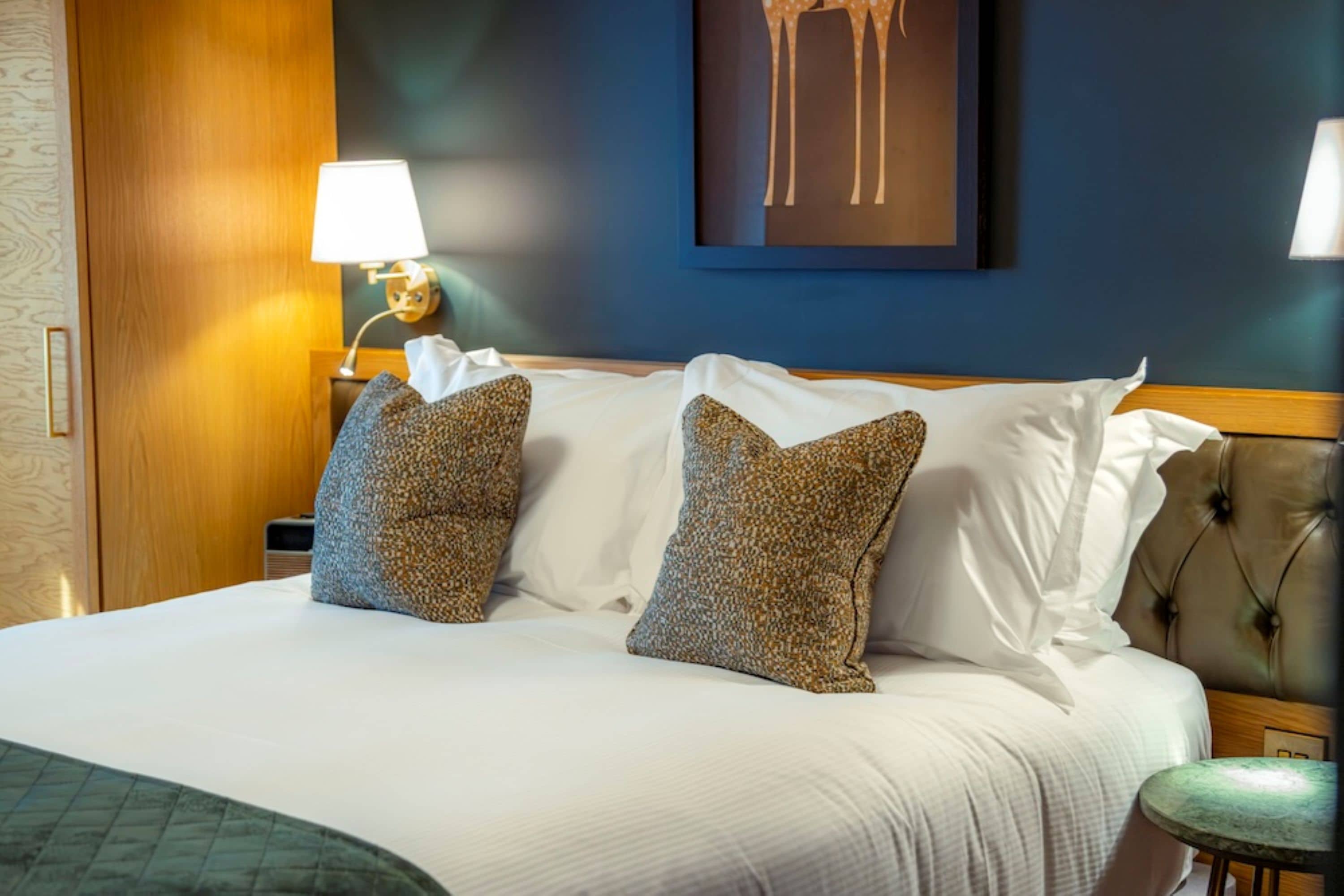 Une chambre d’hôtel avec un lit, des oreillers, une table de chevet, une lampe, une œuvre d’art murale et un fauteuil vert.