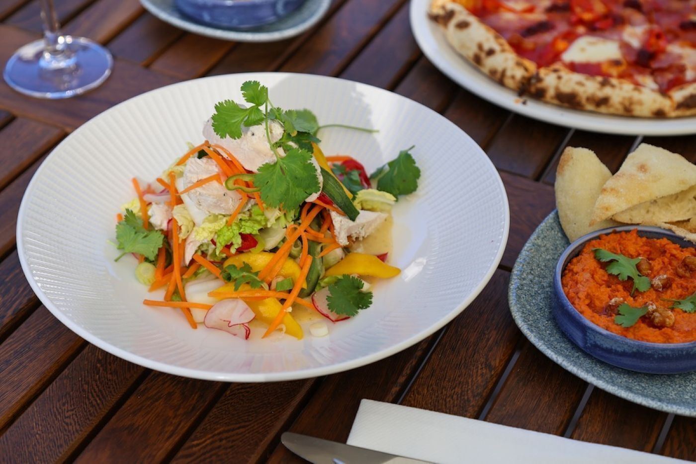 A white plate with a salad, a bowl of red dip, and a pizza on a wooden table.