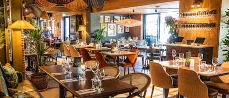 Un restaurante con mesas dispuestas para comer, sillas de mimbre, lámparas colgantes y una zona de bar con vasos.