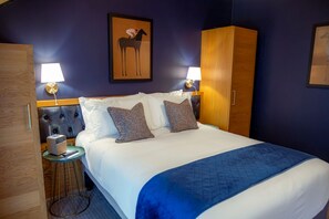 A hotel room with a bed, two bedside lamps, a framed picture on the wall, a wardrobe, and a small table with a lamp. - The Royal Foresters, Ascot, Berkshire - Acorn Pubs (Ascot)