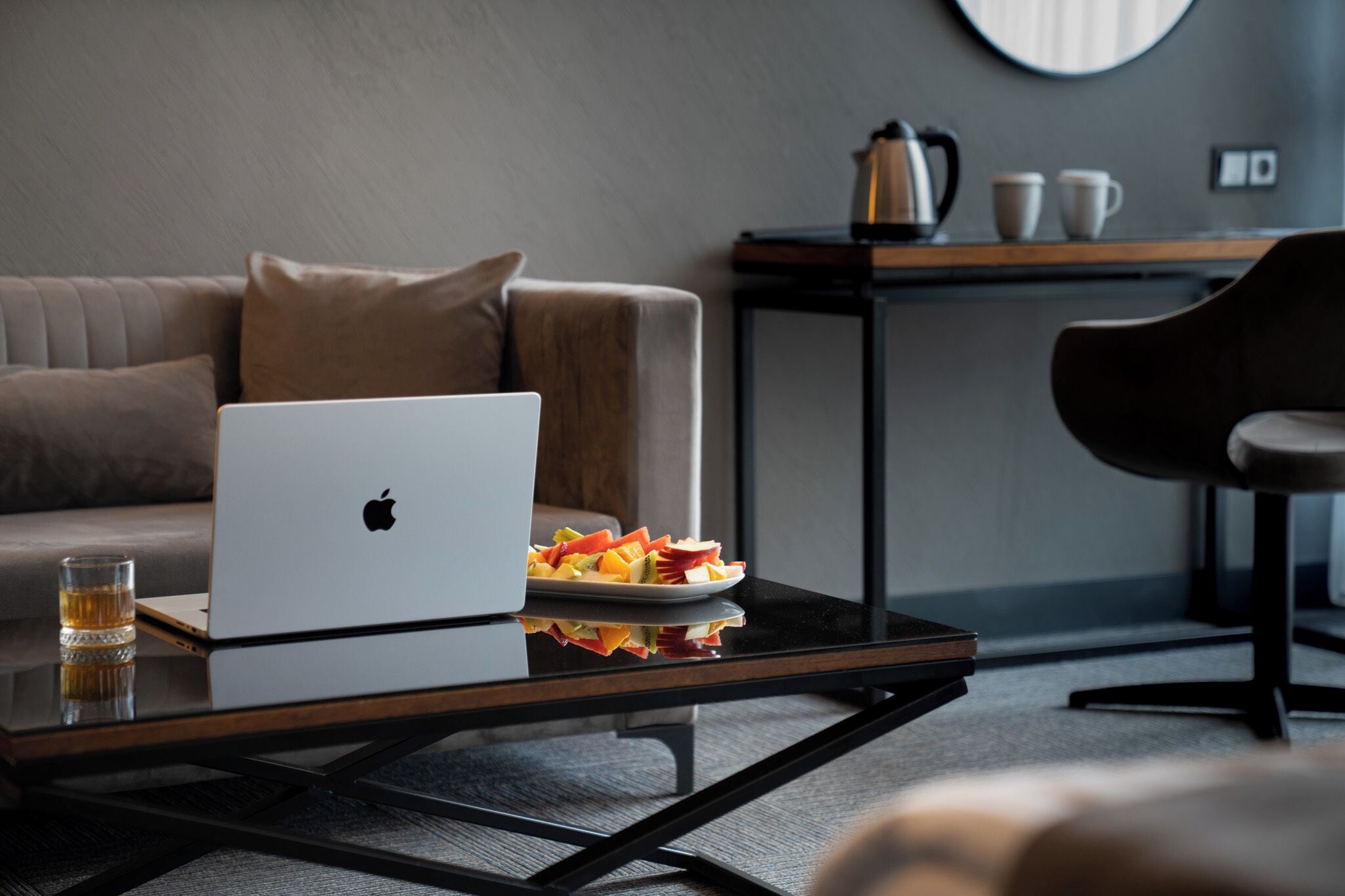 Una habitación moderna con un sofá, una mesa de vidrio con una computadora portátil y fruta, y una mesita con tazas y un hervidor.