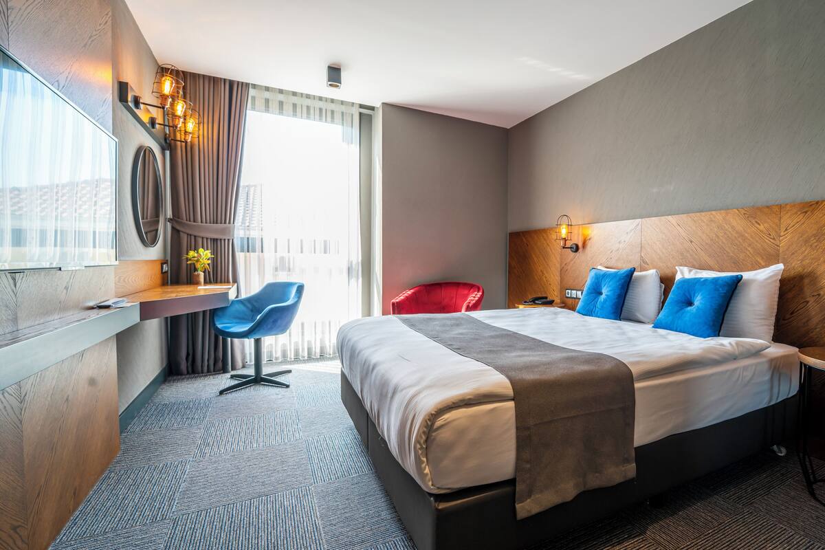 A modern hotel room with a large bed, a desk, a red chair, and a small table with a vase of flowers.