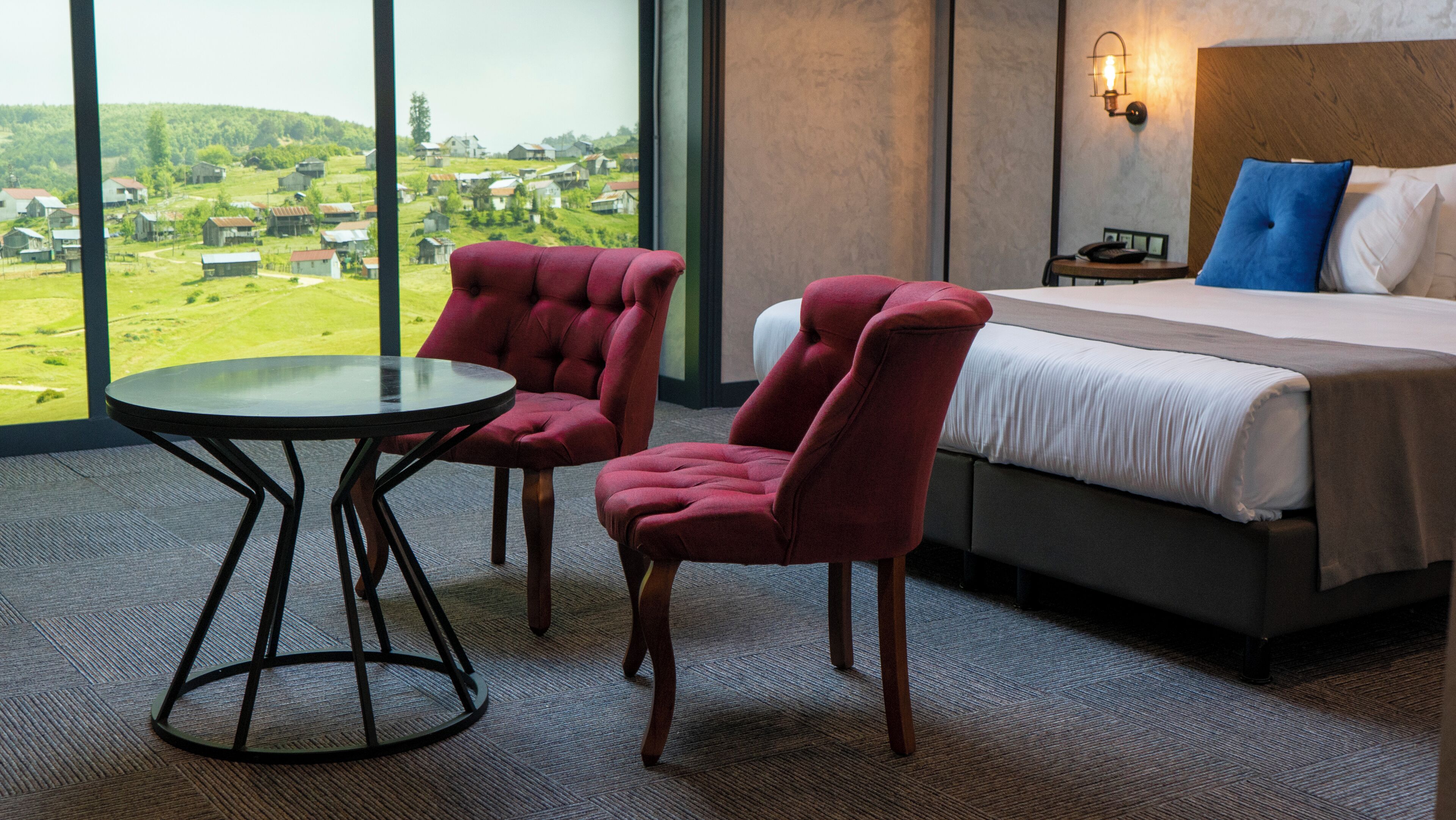 Camera d'albergo con un letto, due poltrone rosse, un tavolino e vista su un paesaggio verde attraverso ampie finestre.