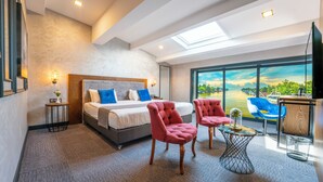 A modern hotel room with a large bed, two red armchairs, a small side table, and a large window with a view of a body of water.