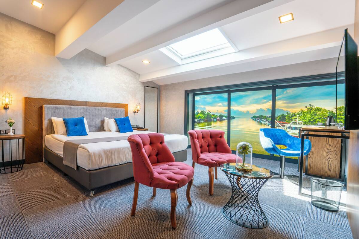 A modern hotel room with a large bed, two red armchairs, a small side table, and a large window with a view of a body of water.