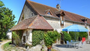 Un bâtiment rustique en pierre, doté d’un toit en tuiles, d’une allée en gravier et d’un patio aménagé avec des sièges d’extérieur.