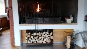 A fireplace with a burning fire, a wooden ceiling, and a stack of firewood below.