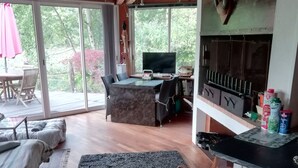 A spacious living area with a wooden ceiling, a dining table with chairs, a sofa, and a TV. There is a grill and a bookshelf with books and decorative items.