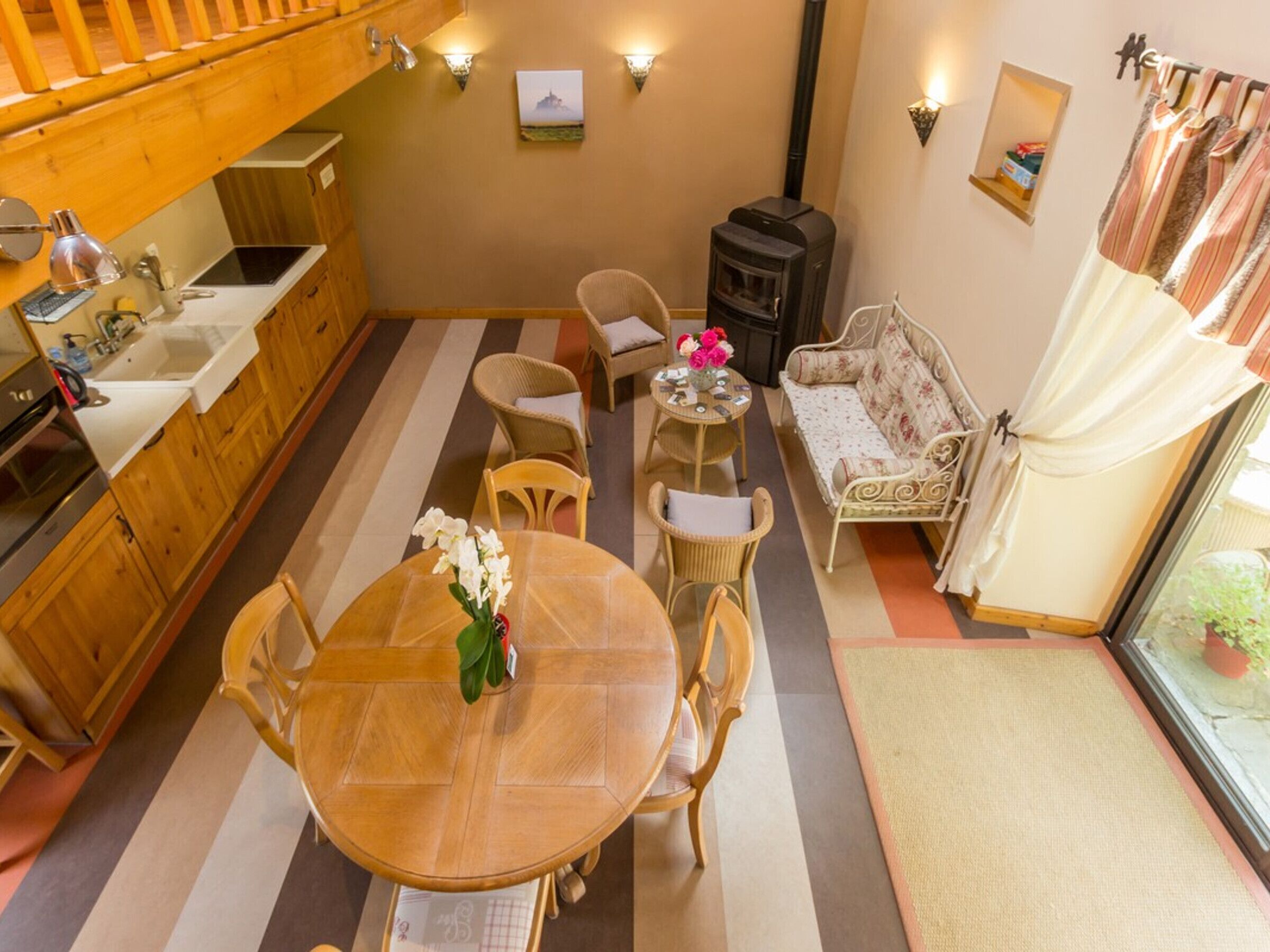 Een gezellige kamer met een houten eettafel, stoelen, een bank, een klein tafeltje met bloemen, een open haard en een keuken met houten kastjes en een spoelbak.