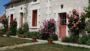 A stone building with a red-tiled roof, multiple windows with wooden shutters, and a variety of potted plants and flowers in front. - Touraine Cottage near Loches with Fireplace and Garden (Saint-Senoch)