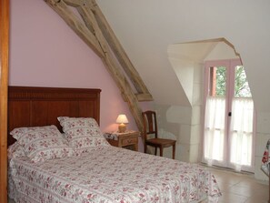 A bedroom with a wooden sloped ceiling, a bed with floral bedding, a nightstand with a lamp, and a chair.