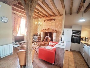 A cozy living room with a fireplace, wooden beams, a red sofa, and a kitchen with modern appliances. - Touraine Cottage near Loches with Fireplace and Garden (Saint-Senoch)