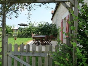 Une porte en bois mène à un patio agrémenté d’une table et de chaises, le tout entouré de verdure et bordé d’un bâtiment aux volets rouges.