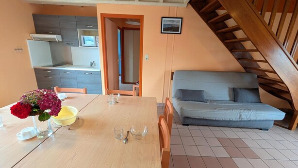 Een eetgedeelte met een houten tafel en stoelen, een keuken op de achtergrond en een trap naar een hoger niveau.