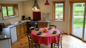 Una cocina compacta con armarios de madera, una mesa de comedor preparada para cuatro comensales y un ventanal con vistas a zonas verdes.