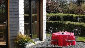 Un edificio con un gran ventanal corredero, una mesa roja con sillas y una planta en maceta.