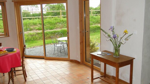 Una habitación con una mesa roja, sillas, una mesita de madera con un jarrón de flores y un ventanal corredero que da a un jardín.