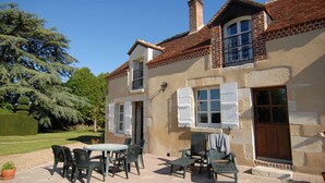 Une maison à deux étages avec une façade en briques, un toit en tuiles et un petit balcon. On y trouve une terrasse avec des meubles d’extérieur et un pot de fleurs.
