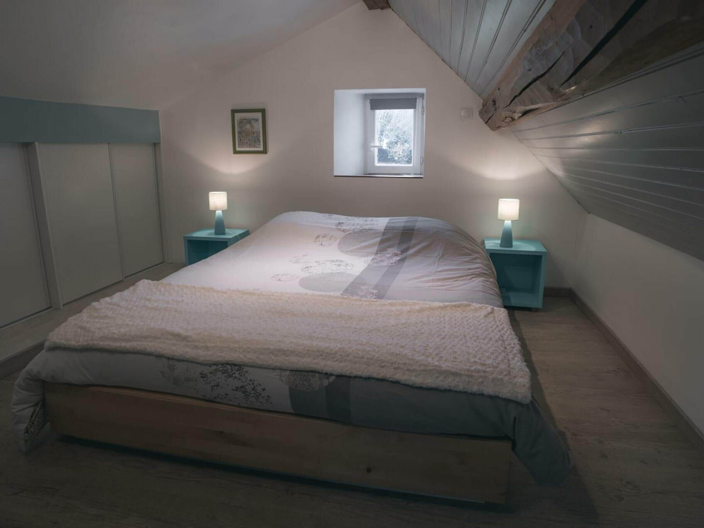 Une chambre avec un plafond en pente, une fenêtre, deux tables de chevet avec des lampes et un lit avec une couverture.