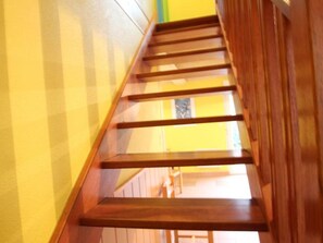 Un escalier en bois avec une rampe, menant à une pièce dont un mur est jaune et qui possède une fenêtre.