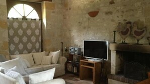 A rustic living room with a sofa, a television on a wooden stand, a fireplace with decorative tiles, and a patterned curtain. - Charming cottage in Chinon forest with private garden (Saint-Benoît-la-Forêt)
