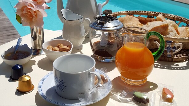 En morgenmadsopstilling med et bord, en tepotte, kopper, en kande, en kurv med brød og en vase med blomster ved en swimmingpool.