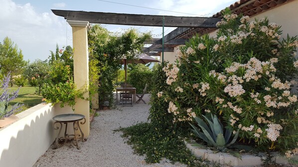 Un espacio de estar al aire libre techado, con una mesa y sillas, rodeado de follaje y plantas con flores.