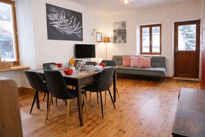 Ruokailutilassa on puinen pöytä, tuoleja ja televisio. Siellä on myös sohva, jossa on koristetyynyjä, sekä ovi, josta on näkymä ulos.