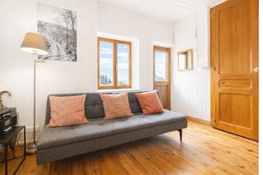 Huoneessa on harmaa sohva, kaksi kuviollista koristetyynyä, lattiavalaisin, taulu ja puiset ovet.