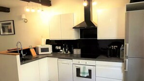 Ett kompakt kök med vita skåp, en svart köksfläkt, en mikrovågsugn, en diskmaskin och ett kylskåp.