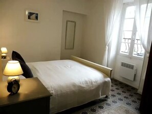 Habitación de hotel con cama, mesita de noche con lámpara y despertador, ventana con cortinas y radiador.