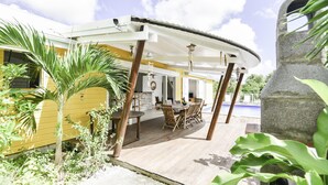 A covered outdoor patio with a pool, wooden deck, and yellow building.