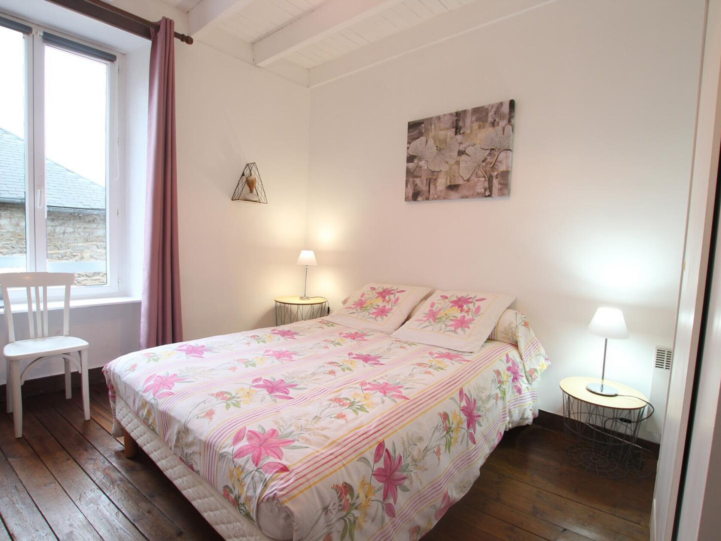 A bedroom with a floral bedspread, a chair by the window, and a small round table with a lamp.