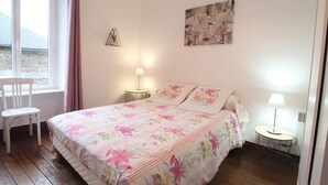 Un dormitorio con cubrecama floral, una silla junto a la ventana y una mesita redonda con una lámpara.
