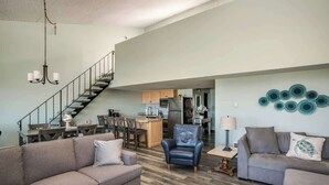 Een ruime woonkamer met een bank, fauteuil en een eettafel. Een trap leidt naar een hoger gelegen verdieping.