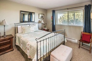 A bedroom with a single bed, a nightstand, a chair, a window with curtains, and a mirror on the wall.