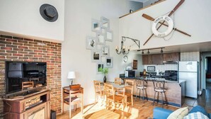Una sala de estar con chimenea de ladrillo, televisor, muebles de madera, comedor y cocina con barra.