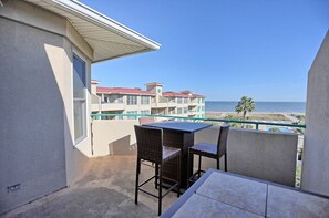 Varanda com mesa e cadeiras, vista para uma praia e um edifício com telhado vermelho.