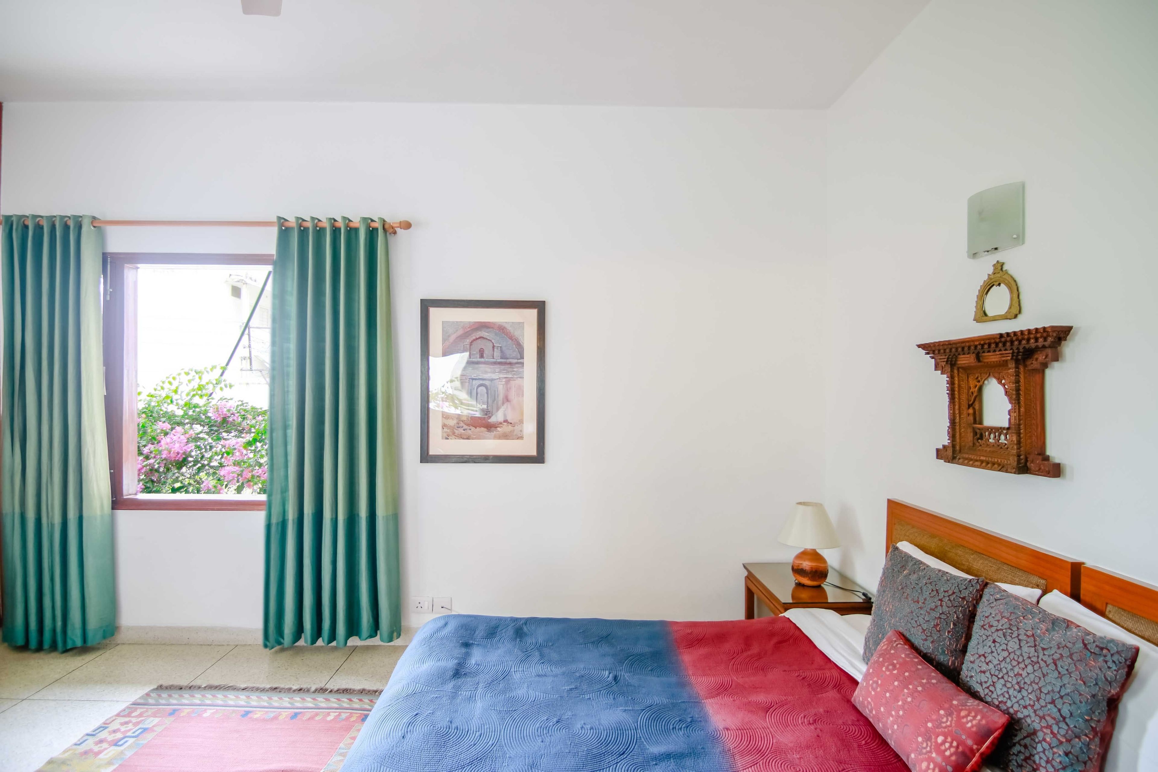 A bedroom with a bed, curtains, a framed picture, a nightstand, and a wall-mounted mirror.
