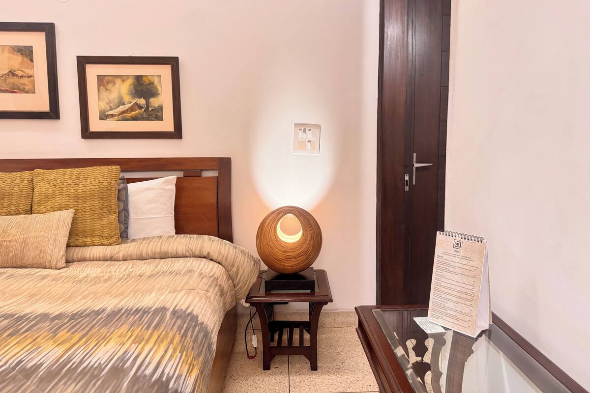 A neatly arranged bedroom with a bed, a bedside lamp, a small table with a menu, and framed artwork on the wall.