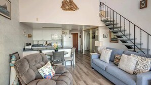 A spacious living area with a grey sofa, a blue sofa with patterned cushions, a kitchen in the background, and a staircase.