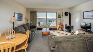 Uma sala de estar com sofá, mesa de jantar com cadeiras, televisão, lareira e uma janela grande com vista.