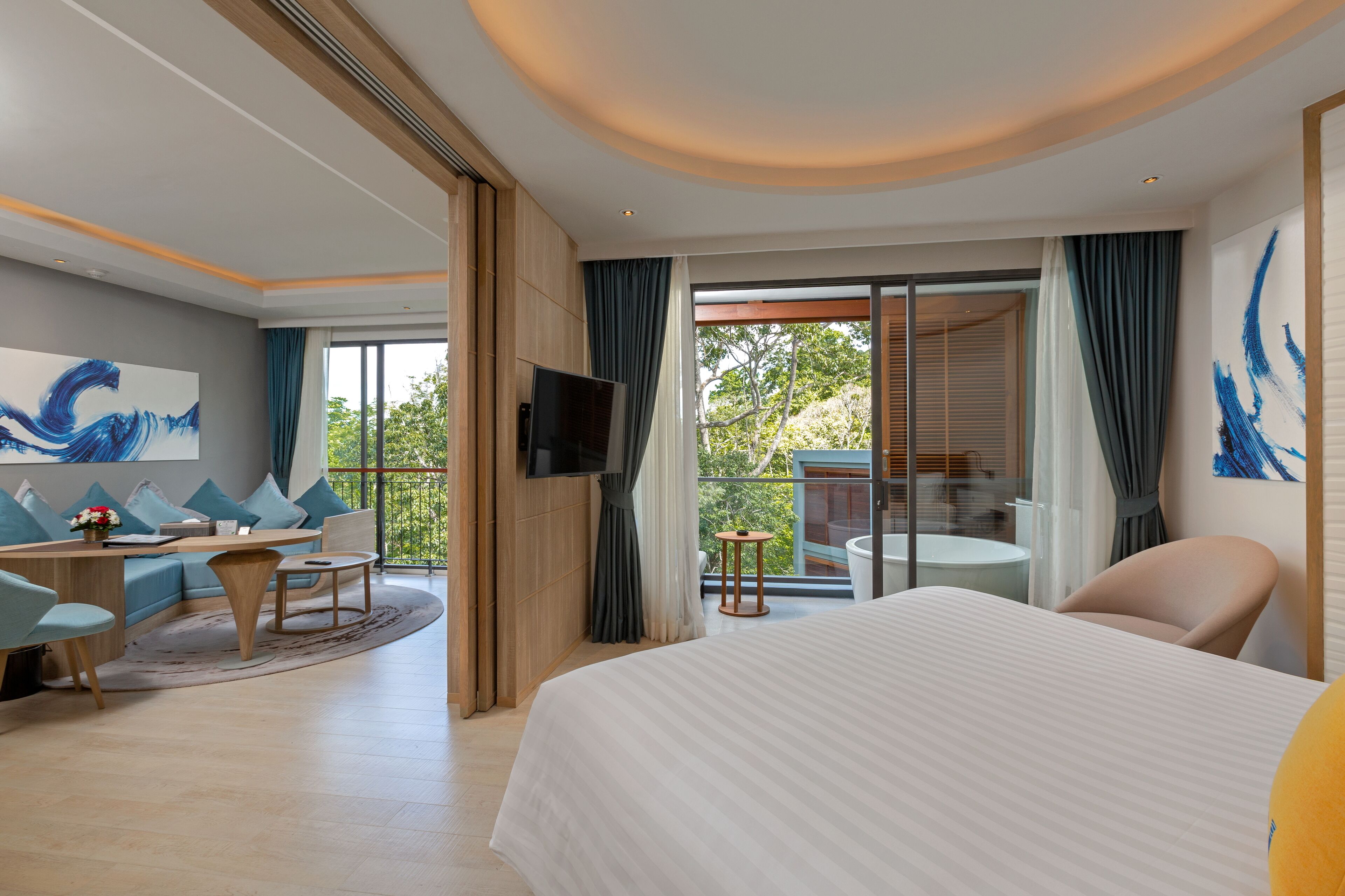 Habitación de hotel moderna con una cama grande, un sofá, un televisor y un balcón con vistas a árboles.