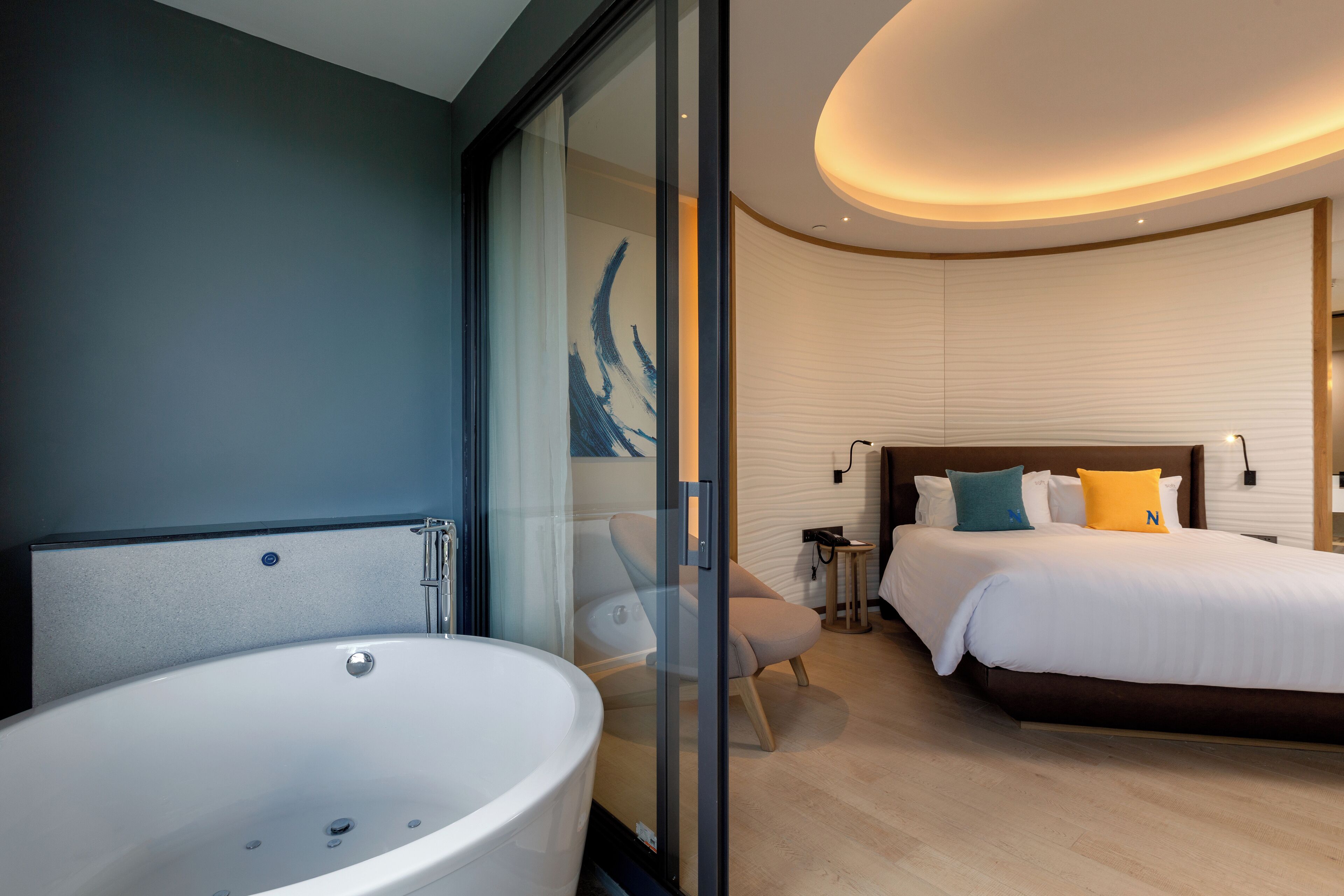 Une chambre d’hôtel moderne dotée d’une grande baignoire, d’un lit bien fait avec des oreillers, et d’une paroi en verre qui sépare la salle de bain de la chambre.