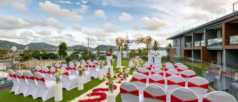 A wedding ceremony setup with rows of white chairs with red sashes, a floral aisle, and a building in the background.