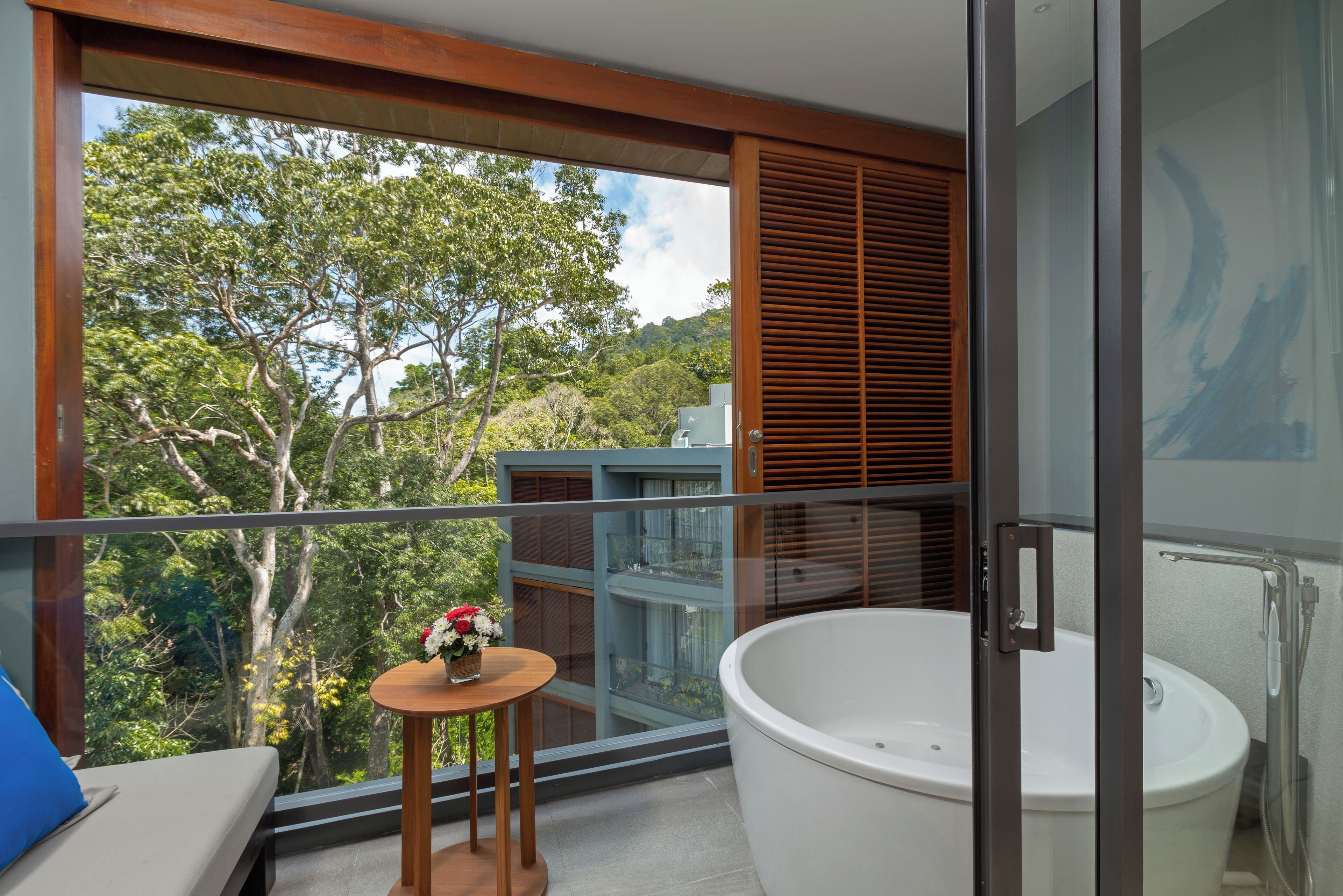 A balcony with a view of trees and a building, a white bathtub, and a small table with flowers.