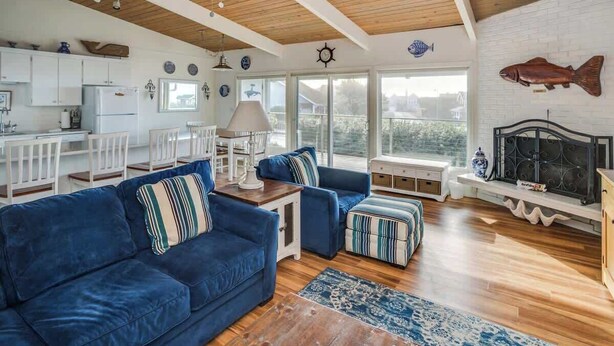A spacious living area with a blue sofa, ottoman, and a fireplace. There is a kitchen with white cabinets and a dining area with white chairs.